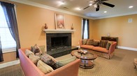 This single-floor space exudes a classic, welcoming ambiance defined by thoughtful architectural details and tasteful finishes. The living area serves as the heart of the space, anchored by a standout fireplace dressed in rich dark granite surround and a crisp white mantel — a focal point that immediately draws the eye. Warm, earthy tones on the walls complement the neutral carpeted flooring, creating a cohesive and comfortable atmosphere throughout. Crown molding adds a refined architectural touch, framing the ceilings with elegant detail. The room is illuminated by a combination of recessed lighting and a ceiling fan fitted with a decorative light fixture, ensuring both functionality and style. Generous seating, including plush coral-toned sofas accented with patterned throw pillows, invites relaxation, while a round glass-top coffee table adds a contemporary contrast to the traditional aesthetic. Natural light filters in through curtained windows, softening the overall feel of the space. A framed still-life artwork displayed above the mantel and decorative ceramic vases complete the polished, curated look of this inviting living environment.