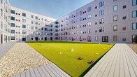 Located at 2171 E 103rd St in Cleveland, Ohio, this outdoor space is defined by its modern architectural setting and thoughtfully designed open-air environment. The space is framed on multiple sides by multi-story buildings clad in light gray metal panel siding, punctuated by an asymmetric arrangement of windows that lend a contemporary, geometric aesthetic to the complex.

At the heart of the space lies a large, open courtyard surfaced with vibrant artificial turf, providing a clean, low-maintenance green area. Bordering the turf is a wide composite wood deck, offering a transitional surface between the building entrances and the central green space. Glass double doors open directly onto the deck, providing convenient access to and from the building interiors.

Surrounding portions of the deck, gravel landscaping adds textural contrast and aids in drainage. A metal railing system lines the perimeter of an elevated walkway, offering open views of the surrounding Cleveland neighborhood, including mature trees and the broader urban landscape beyond.

The space benefits from abundant natural light and open sky views, creating a bright and welcoming atmosphere. The overall design balances functionality with a clean, contemporary aesthetic well-suited to its urban setting.