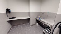 This single-floor space is purpose-built for pet washing or grooming, combining practicality with a clean, utilitarian aesthetic. The room features large-format gray tile flooring that provides durability and ease of cleaning, while the walls are finished with a two-tone treatment — gray tile wainscoting on the lower half and smooth white paint above — creating a hygienic and visually organized environment. A stainless steel wash tub with dedicated plumbing, including visible supply lines and a hose attachment, is positioned along one wall, offering a fully functional bathing station. A protective side panel flanks the tub, helping to contain splashing during use. Opposite the wash station, a built-in bench with a speckled terrazzo-style surface provides a convenient spot for preparation or storage, supported by an open base below. The room's neutral color palette of grays and whites contributes to a clean, professional atmosphere. The space is efficiently laid out to maximize utility within a compact footprint, making it well-suited for grooming operations in a residential building amenity, veterinary, or pet care facility context.