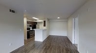 This single-floor space features an open-concept design that seamlessly connects the kitchen and living area, creating a bright and functional environment. The flooring throughout is a rich, dark hardwood-style surface that adds warmth and contrast to the crisp white walls and light ceiling.

The kitchen is well-appointed with dark espresso-toned cabinetry that extends to the upper walls, providing ample storage. Stainless steel appliances, including a gas range and refrigerator, lend a modern edge to the space. A peninsula with a light-colored countertop extends into the open area, offering additional prep space and serving as a natural divider between the kitchen and the adjacent living zone. Recessed and overhead lighting ensures the kitchen remains well-lit.

The living area is generously proportioned, offering an open and versatile space suitable for a variety of furniture configurations. Neutral white walls provide a clean backdrop, while recessed lighting maintains consistent illumination. A door on the far side of the living area hints at additional access or an adjoining room, contributing to the overall functional flow of the layout.