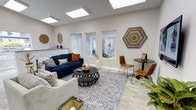 This single-floor space is thoughtfully designed to balance comfort and functionality. The main living area is immediately welcoming, bathed in abundant natural light streaming through multiple windows and a set of overhead skylights, which enhance the open, airy feel of the room. Light-toned walls and light wood-style flooring contribute to the bright, clean aesthetic throughout.

The seating area is anchored by a navy blue sofa and a complementary cream-colored couch, arranged around a dark, lattice-style round coffee table atop a patterned area rug. A small bistro-style table flanked by two mustard-colored chairs offers a secondary seating option, ideal for informal conversations or small group gatherings. Decorative touches, including a large hexagonal wall art piece, a round wall clock, and carefully placed greenery, add warmth and personality to the space.

A compact service or storage area is visible in the background, featuring white cabinetry that provides practical functionality without disrupting the overall aesthetic. The combination of skylights, thoughtful décor, and versatile furniture arrangements makes this a well-rounded, inviting space suited for a variety of uses.