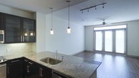 This single-floor space showcases a thoughtfully designed open-concept layout that seamlessly connects the kitchen and living areas. The kitchen is anchored by a substantial island featuring granite countertops with attractive veining, a stainless steel sink, and dark cabinetry that provides a striking contrast to the light-toned walls. Two contemporary pendant lights with frosted glass shades hang above the island, complementing a track lighting system overhead that allows for flexible illumination throughout the space.

The living area extends naturally from the kitchen, finished with warm hardwood flooring that runs throughout the entire space, contributing to a cohesive and polished aesthetic. A ceiling fan is centrally positioned in the living area, enhancing both comfort and airflow. Natural light enters generously through a set of glass French doors at the far end of the room, fitted with horizontal blinds for privacy control. The clean white walls and open floor plan create a bright, airy atmosphere, making the space feel expansive and welcoming. The overall design balances modern finishes with functional details.