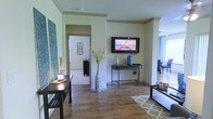 This single-floor space showcases a well-appointed interior defined by warm hardwood flooring that flows seamlessly throughout the open layout. The living area is anchored by a wall-mounted flat-screen television positioned above a sleek dark console table, which is styled with decorative vases and accent pieces. Crown molding along the ceiling adds a refined architectural detail, lending the space a polished, finished look. Light-toned walls in a soft sage-green hue create a calm, neutral backdrop that complements the rich wood flooring below. A tall, textured floor vase with yellow floral branches adds a bold decorative focal point near the room's central structural column, which subtly defines the space without closing it off. To the left, a dark wooden side table with a table lamp and wall art provides additional character. A doorway visible from the main living area leads to an adjacent room, hinting at a connected, functional floor plan. To the right, an adjoining area with additional seating and hardwood flooring extends the living space further. A geometric-patterned area rug grounds the seating arrangement, adding texture and visual interest. Overall, the space presents a cohesive, contemporary aesthetic with warm tones and thoughtful detailing throughout.
