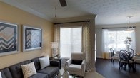 This single-story space showcases a thoughtfully designed living area that balances comfort and style. The living room is highlighted by warm, golden-toned walls that create an inviting ambiance, complemented by a textured ceiling with crown molding and a ceiling fan that adds both function and visual interest. Large windows dressed with patterned drapery panels allow generous amounts of natural light to fill the room, enhancing the bright and airy feel of the space. A second window visible in the background further reinforces the well-lit character of the interior. The living area is furnished with a dark upholstered sofa accented with decorative pillows and a chevron-patterned armchair, creating a cohesive and stylish seating arrangement. A side table with a classic lamp and decorative vases on the coffee table add refined finishing touches. Dark hardwood-style flooring anchors the space and contrasts elegantly with the lighter wall tones. The overall ambiance is warm, polished, and welcoming, offering a well-curated environment that feels both livable and aesthetically considered.