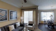 This single-story space showcases a thoughtfully designed living area that balances comfort and style. The living room is highlighted by warm, golden-toned walls that create an inviting ambiance, complemented by a textured ceiling with crown molding and a ceiling fan that adds both function and visual interest. Large windows dressed with patterned drapery panels allow generous amounts of natural light to fill the room, enhancing the bright and airy feel of the space. A second window visible in the background further reinforces the well-lit character of the interior. The living area is furnished with a dark upholstered sofa accented with decorative pillows and a chevron-patterned armchair, creating a cohesive and stylish seating arrangement. A side table with a classic lamp and decorative vases on the coffee table add refined finishing touches. Dark hardwood-style flooring anchors the space and contrasts elegantly with the lighter wall tones. The overall ambiance is warm, polished, and welcoming, offering a well-curated environment that feels both livable and aesthetically considered.