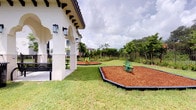This single-story outdoor amenity space showcases a thoughtfully designed recreational area that blends architectural elegance with functional play facilities. The building adjacent to the grounds features a distinctive Mediterranean-inspired design, characterized by white stucco walls, arched colonnades, dark wood bracket accents beneath a clay-tiled roofline, and wall-mounted lantern-style light fixtures that add a classic aesthetic appeal.

The grounds are landscaped with well-maintained open lawn areas that provide generous open space for leisure. Two dedicated play zones are positioned throughout the yard, each edged with bordered mulch beds filled with wood chip surfacing for safety. Colorful green play equipment, including a spring rider and a circular climber, offers recreational options for children. A third, smaller circular play pad is positioned further along the space, adding depth and variety to the layout.

Seating is available along the covered arcade via classic metal bench seating, offering a shaded area for observation and rest. The entire perimeter is enclosed by a black iron fence, ensuring security while maintaining visual openness to the surrounding greenery and mature tropical landscaping beyond.