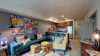This single-floor residence makes a bold first impression with its eclectic, art-forward interior design. The open-concept living and dining area serves as the heart of the home, anchored by a striking large-scale pop-art canvas that dominates one wall, setting an energetic and creative tone throughout. The living space is generously proportioned, featuring light wood flooring, recessed lighting, and a vibrant mix of teal and yellow furnishings layered over a colorful area rug. A large window bathes the seating area in natural light while offering views of the surrounding building exterior. The kitchen flows seamlessly from the living area and is equipped with warm wood cabinetry, granite countertops, a tiled backsplash, and a full suite of stainless steel appliances including a refrigerator, range, and microwave. A breakfast bar with seating provides a casual dining option. The bedroom is a well-balanced retreat, offering a walk-in closet, a dedicated workspace with a desk and mirror, and a cozy reading nook by the window. Bold artwork and richly colored textiles continue the home's artistic theme. The bathroom features granite countertops, wood-toned cabinetry, and light wood flooring, with a decorative sunburst mirror adding a stylish accent. Throughout, the space blends contemporary finishes with expressive, gallery-worthy décor.