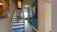 This two-floor residence makes a strong first impression with its thoughtfully designed entryway, where rich dark hardwood-style flooring sets the tone for the interior. A bold teal accent wall, adorned with framed wall art, adds a striking pop of color that contrasts beautifully with the neutral tones throughout the space. A carpeted staircase with a light wood banister connects the two levels, offering both functionality and visual interest from the moment you enter.

From the entry, sightlines open toward a bright and airy living area, where abundant natural light pours in through large windows, illuminating the space and creating an inviting atmosphere. A ceiling fan is visible overhead, contributing to the comfortable, lived-in feel of the main level. Adjacent to the living area, the kitchen is equipped with stainless steel appliances and features light cabinetry, offering a clean and modern aesthetic that complements the overall design of the home.

The open layout of the main floor encourages a natural flow between the entryway, kitchen, and living spaces, making the home well-suited for both everyday living and entertaining. The combination of warm accent colors, modern finishes, and plentiful natural light throughout gives the space a balanced and welcoming character across both floors.