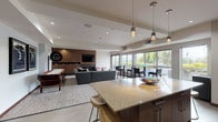 Located at 3261 SW Avalon Way in Seattle's West Seattle neighborhood, this single-floor amenity space offers a thoughtfully designed open-plan layout that seamlessly integrates a full kitchen, social lounge, and entertainment area. The kitchen is a standout feature, anchored by a generously sized granite-topped island with seating for multiple guests, complemented by rich walnut cabinetry, dark stone countertops, a light blue glass tile backsplash, stainless steel appliances including a French door refrigerator, and pendant lighting overhead. The open living and lounge area extends naturally from the kitchen, featuring plush sectional seating, a large flat-screen TV mounted within a floor-to-ceiling walnut media wall, and a sleek built-in fireplace below. A shuffleboard table and wall-mounted billiards sign add a playful, recreational dimension to the space. Recessed lighting and tray ceilings contribute to a polished, modern aesthetic throughout. Large windows and glass sliding doors flood the space with natural light and offer views of the surrounding greenery, with access to an adjacent outdoor terrace. The overall ambiance is contemporary and inviting, making it well-suited for gatherings, casual socializing, or relaxed recreation.