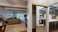 This single-floor residence showcases a cohesive design defined by distinctive exposed beam ceilings with textured finishes found throughout the main living areas. The living room is a comfortable, well-proportioned space with plush carpeting and large sliding glass doors that open onto a private balcony, offering pleasant outdoor views of surrounding trees. Vertical blinds provide flexible light control, while a gold-framed oval mirror and wall sconces add a classic decorative touch.

Adjacent to the living room, the dining area features wood-look flooring, a ceiling fan with integrated lighting, and ample space for a full dining set. The kitchen is efficiently laid out with dark wood cabinetry, white appliances including a range and refrigerator, and light countertops. A mirrored sliding closet door near the kitchen entry reflects the space and adds visual depth.

The bedroom is a serene retreat with soft carpeting, exposed beam ceilings, and access to a walk-in closet. The full bathroom includes a tub-shower combination, wood-look flooring, a white vanity, and textured walls. Throughout the residence, a warm, traditional ambiance is maintained by consistent finishes and thoughtful details.