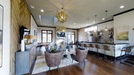 This well-appointed single-floor space exudes a refined, contemporary aesthetic that seamlessly blends comfort with style. The main lounge area is anchored by a stunning floor-to-ceiling stone fireplace, above which a flat-screen television is mounted, creating a natural focal point for the room. Rich, dark hardwood flooring runs throughout, complementing the warm, neutral color palette of the furnishings.

Seating is plentiful and thoughtfully arranged, featuring plush upholstered lounge chairs and sofas grouped around a central area rug, ideal for relaxed conversation or gathering. A decorative floral arrangement adds a touch of organic elegance to the space.

An adjacent bar and kitchen area offers dark countertop surfaces with high-top bar seating, creating a social hub perfect for entertaining. A refrigerator and open shelving complete this functional zone.

Overhead, eye-catching gold orb pendant chandeliers serve as artistic focal points, while recessed lighting ensures the space is well-lit throughout. A skylight further enhances the bright, airy atmosphere. Large windows with warm wood trim flood the interior with natural light and offer views to the outdoors. The overall ambiance is inviting and upscale, making this a distinguished space for socializing and relaxation.