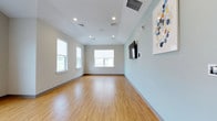 This single-floor space offers a clean, modern design suited for a variety of uses. The open layout provides generous room to move freely, with warm hardwood-style flooring running throughout, adding both visual warmth and durability to the space.

The walls are finished in soft, calming tones — a warm beige in some areas and a cool light aqua in others — creating a welcoming and pleasant atmosphere. Recessed ceiling lighting is distributed evenly throughout, ensuring consistent illumination across the entire area. Multiple windows along the perimeter walls allow abundant natural light to fill the space, offering views of the surrounding exterior environment.

A large wall-mounted flat-screen television is positioned prominently on one of the aqua-toned accent walls, alongside a framed abstract artwork featuring soft blue, gold, and neutral tones, adding a decorative touch. Smart panel controls are also mounted nearby. The space includes a doorway leading to an adjacent area, suggesting additional rooms or facilities beyond the main open area.

The overall ambiance is bright, airy, and versatile, with a contemporary aesthetic that balances functionality with a clean, organized design sensibility.