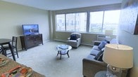 This single-floor residence centers around a well-appointed living room designed for both comfort and functionality. The space is finished with neutral-toned carpeting and warm beige walls, creating a calm and inviting atmosphere throughout.

The living room is notably bright, benefiting from a large bank of windows that span a significant portion of one wall. These windows frame sweeping views of the surrounding cityscape and green treetops below, allowing generous natural light to fill the room. A wall-mounted air conditioning unit is positioned beneath the windows, complementing the baseboard heating system that runs along the perimeter.

The room is furnished with a plush gray sofa accented by decorative pillows, a classic checkered armchair, and a round glass-top coffee table with a curved dark wood base. A dark wood media console anchors the opposite wall, supporting a flat-screen television. The layout encourages comfortable conversation and relaxed viewing.

Overall, the space presents a functional and well-lit living environment with a traditional aesthetic, benefiting greatly from its elevated position and the expansive outdoor views it captures.