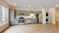 This single-floor unit offers a thoughtfully designed open-concept layout that seamlessly connects the kitchen and living areas. The kitchen is a standout feature, showcasing sage green shaker-style cabinetry extending to the upper walls for generous storage, complemented by granite-look countertops and a classic white subway tile backsplash. A full suite of stainless steel appliances — including a gas range, built-in microwave, dishwasher, and refrigerator — provides everything needed for everyday cooking. A matte black faucet at the double sink adds a modern accent. Light luxury vinyl plank flooring runs throughout, lending warmth and continuity to the space. The living area is generously sized, bathed in natural light from large windows that frame views of the surrounding neighborhood. A wall-mounted HVAC unit ensures year-round comfort. The bathroom is finished in a clean, contemporary style with white surfaces, shaker-style vanity cabinetry with matte black hardware, a wide vanity mirror, and a full tub-shower combination. Recessed lighting throughout the unit provides consistent, well-distributed illumination, contributing to the overall bright and modern ambiance of this move-in-ready space.