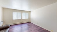 This single-floor, 3-bedroom, 1-bathroom unit offers a practical and well-organized layout across a compact footprint. The living room serves as the welcoming centerpiece of the space, featuring rich, dark hardwood-style flooring, neutral cream-toned walls, and dual windows fitted with horizontal blinds that allow natural light to filter in. Baseboard heating runs along the walls, ensuring consistent warmth throughout. The kitchen is conveniently open to the living area and is equipped with warm wood cabinetry, ample upper and lower storage, a white refrigerator, a sink with countertop workspace, and overhead lighting — offering a functional cooking environment. The three bedrooms are carpeted in a soft grey tone, providing a comfortable feel underfoot. Each bedroom includes at least one window with blinds and baseboard heating. The primary bedroom offers a generous amount of space, while the additional bedrooms are well-suited for various uses. A utility access panel is notably present in one of the bedrooms. The shared full bathroom features a tub and shower combo, a wood-framed vanity with a white sink, a mirrored medicine cabinet, and track lighting above. The overall aesthetic is clean, neutral, and functional, making this a straightforward and livable space.