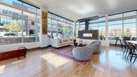 This expansive lounge is a thoughtfully designed communal space that seamlessly blends comfort, entertainment, and modern aesthetics across a single open floor plan. Floor-to-ceiling glass windows line multiple walls, flooding the interior with abundant natural light and offering unobstructed views of the surrounding streetscape and greenery.

The main seating area anchors the space with a generous sectional sofa, rounded accent chairs, and a vibrant Persian-style area rug, all positioned around a sleek brick-clad fireplace that serves as a striking focal point. Natural hardwood flooring flows continuously throughout, unifying the various activity zones.

A dedicated game area features a full-size billiards table beneath geometric brass pendant lights, while an adjacent kitchen and bar area provides a long live-edge communal table with bar seating, stainless steel appliances, a wall-mounted TV, and a textured white stone accent wall.

A more intimate lounge nook offers a cozy retreat, distinguished by sculptural green wave-textured walls, warm globe pendant lighting, a leather banquette, and compact café-style tables. Exposed brick columns and curated artwork throughout add visual depth and character, creating an overall atmosphere that is social, stylish, and inviting.