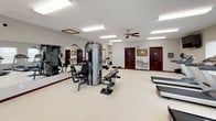This fitness center offers a spacious, single-floor layout designed to accommodate a comprehensive workout experience. The open floor plan is finished with durable speckled flooring throughout, providing a clean and functional surface for exercise.

The cardio section is well-appointed with multiple treadmills and elliptical machines, giving users a variety of options for aerobic training. A wall-mounted flat-screen television is positioned for easy viewing during workouts. Ceiling fans provide additional air circulation, complementing the bright fluorescent lighting overhead.

The strength training area features a multi-station cable machine along with dedicated workout benches, catering to a range of resistance exercises. A large wall-length mirror allows users to monitor their form, while a dumbbell rack provides free weight options. A bulletin board is mounted near the entrance for notices and informational postings.

Natural light fills the space through multiple windows fitted with white blinds, offering views of the surrounding outdoor landscape. A built-in water fountain is conveniently located near the entrance, and a security camera ensures a safe environment. The facility includes clearly marked emergency exits and fire safety equipment, reflecting a strong commitment to user safety and comfort.