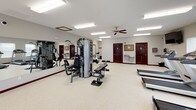 This fitness center offers a spacious, single-floor layout designed to accommodate a comprehensive workout experience. The open floor plan is finished with durable speckled flooring throughout, providing a clean and functional surface for exercise.

The cardio section is well-appointed with multiple treadmills and elliptical machines, giving users a variety of options for aerobic training. A wall-mounted flat-screen television is positioned for easy viewing during workouts. Ceiling fans provide additional air circulation, complementing the bright fluorescent lighting overhead.

The strength training area features a multi-station cable machine along with dedicated workout benches, catering to a range of resistance exercises. A large wall-length mirror allows users to monitor their form, while a dumbbell rack provides free weight options. A bulletin board is mounted near the entrance for notices and informational postings.

Natural light fills the space through multiple windows fitted with white blinds, offering views of the surrounding outdoor landscape. A built-in water fountain is conveniently located near the entrance, and a security camera ensures a safe environment. The facility includes clearly marked emergency exits and fire safety equipment, reflecting a strong commitment to user safety and comfort.
