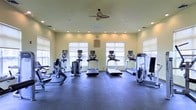 This single-story fitness center offers a clean, functional workout environment designed to accommodate a range of cardiovascular exercise needs. The room is outfitted with multiple treadmills and elliptical-style machines, each equipped with individual digital display screens, arranged in a row facing large windows that allow generous natural light to fill the space. Roller shades on each window provide adjustable light control throughout the day. The flooring consists of durable dark rubber matting, well-suited for high-traffic fitness use and providing cushioning underfoot. Recessed lighting is distributed evenly across the ceiling, supplemented by a central ceiling fan that aids in air circulation during workouts. The walls are finished in a soft yellow-green tone, contributing to a bright and energizing atmosphere. Additional strength or resistance equipment is positioned along the sides of the room, maximizing the use of the available floor space. A framed notice or informational board is mounted on the wall between the windows, adding a practical element to the space. Overall, the fitness center presents a well-organized, inviting environment suited for daily exercise routines.