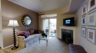 This single-floor residence offers a thoughtfully designed living area that balances comfort and style. The living room serves as the centerpiece of the space, anchored by a classic white-mantled fireplace with slate tile surround, providing both a functional and decorative focal point. A wall-mounted flat-screen television sits above the fireplace, integrating modern entertainment seamlessly into the traditional design.

The room features plush carpeting underfoot and a combination of warm yellow and rich brown accent walls that create a welcoming, layered ambiance. Natural light flows generously through sliding glass doors dressed with elegant patterned drapes, opening directly to a private outdoor patio furnished with a bistro-style table and chairs — ideal for outdoor dining or relaxation.

A decorative sunburst mirror adorns the deep brown feature wall, adding visual depth and a contemporary touch. The furnishings include a generously sized sofa accented with colorful throw pillows, a sleek glass-top coffee table, and additional seating, all arranged to encourage comfortable gathering. The overall aesthetic is warm and inviting, blending classic architectural details with modern accents for a refined yet livable atmosphere.