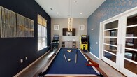 Located at 20220 Bellaire Blvd in Richmond, TX, this single-floor game room is a thoughtfully designed entertainment space that blends modern aesthetics with a fun, social atmosphere. The room is anchored by a full-size pool table with a blue felt surface, positioned beneath a striking cascading chandelier that serves as a dramatic focal point. The interior design features bold navy accent walls paired with eye-catching gold-patterned wallpaper, creating a dynamic and contemporary feel throughout. Multiple large windows allow generous natural light to fill the space, offering views of the surrounding neighborhood. Two classic arcade cabinets — including an Arcade Legends multi-game machine and a Pixel arcade unit — add a retro gaming element that complements the room's lively character. A wall-mounted flat-screen TV and a sleek media console provide additional entertainment options. A built-in bench seat upholstered in blue fabric lines one wall, offering comfortable seating for spectators. Framed artwork and decorative wall hangings add curated visual interest. Elegant French doors with glass panels connect this space to an adjacent room, enhancing the open and accessible layout. The combination of entertainment amenities, stylish finishes, and ample natural light makes this a welcoming and well-appointed community gathering space.