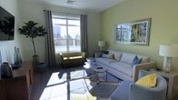 This single-floor space showcases a thoughtfully designed living area that balances comfort and modern style. The sitting room is anchored by a large picture window dressed with patterned grommet curtains, allowing generous natural light to fill the room and offering views of the surrounding outdoor scenery. The walls are finished in a soft, muted green tone that lends a calm and refreshing ambiance throughout.

The living area is furnished with a neutral, light-toned sofa accented with blue and yellow throw pillows, complemented by a chartreuse accent chair that adds a vibrant pop of color. A geometric-patterned area rug in shades of grey, yellow, and white grounds the seating arrangement, while a set of sleek, metallic hexagonal coffee tables serves as a modern centerpiece. A framed nature-inspired artwork adorns one wall, reinforcing the fresh, airy aesthetic of the space.

Dark hardwood flooring runs throughout, providing a rich contrast to the light walls and furnishings. Recessed ceiling vents and well-placed lighting ensure year-round comfort. The overall design is cohesive and contemporary, making this a functional and visually appealing space ideal for relaxing or entertaining.