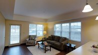 This single-floor space offers a comfortable and well-lit living environment designed for everyday functionality. The living room serves as the centerpiece of the space, immediately drawing attention with its generous natural light streaming through multiple large windows dressed with white horizontal blinds. The warm yellow-toned walls create a cheerful and inviting ambiance, complemented by rich dark hardwood flooring that runs throughout the area and adds a touch of warmth and contrast.

The living room is thoughtfully arranged to maximize both comfort and flow, featuring a spacious seating area with a taupe sofa, a coordinating patterned accent armchair, and a dark wood coffee table styled with books and greenery. A side lamp provides additional warm lighting, while a neatly placed area rug anchors the seating arrangement on top of the hardwood floor. The room's clean white ceiling enhances the sense of openness, and the multiple windows on adjacent walls allow for cross-lighting that keeps the space feeling airy throughout the day. Overall, the space presents a cozy yet functional layout that balances natural light, warm color tones, and classic furnishings seamlessly.