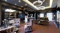 Located at 100 Park Lane Blvd in Cherry Hill, New Jersey, this single-floor space exudes a refined, contemporary aesthetic designed to impress. The interior is anchored by a stunning chandelier arrangement of interlocking circular crystal rings that serve as a dramatic focal point overhead, casting a warm, ambient glow throughout the room. Dark, sophisticated wall tones and recessed lighting complement the space's upscale atmosphere.

The lounge area features elegantly patterned armchairs with bold geometric upholstery, arranged atop a richly colored red and black carpet that adds depth and energy to the room. A sleek, metallic console table with mirrored accents reflects light and enhances the sense of spaciousness.

A built-in bar or service counter with a crisp white surface and dark cabinetry provides a functional yet stylish centerpiece, complete with pendant lighting above and a mounted display screen — ideal for hosting and entertaining. Large windows along one wall invite natural light, offering views to the exterior and balancing the room's darker design palette.

The overall ambiance is polished and modern, combining luxurious finishes, curated furnishings, and thoughtful lighting design to create a versatile and visually striking environment suited for gatherings, social events, or upscale communal use.