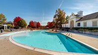 Located at 6400 Preserve Crossing Blvd N in Gahanna, Ohio, this two-story community amenity center offers an impressive array of outdoor recreational features set within a well-maintained residential community. The centerpiece of the outdoor space is a generously sized swimming pool with a gradual entry, complemented by a second pool area, both framed by clean concrete surrounds and classic wrought iron fencing. The expansive sundeck is furnished with rows of modern lounge chairs and shaded umbrella stations, providing ample space for relaxation under open skies. A dedicated sports court area, enclosed by chain-link fencing, adds to the recreational offerings alongside ornamental landscaping with native grasses and vibrant seasonal trees. The two-story clubhouse building features a traditional architectural style with white siding, a gabled entryway with a decorative arch, and an upper-level outdoor terrace outfitted with dining tables and chairs — ideal for social gatherings. Classic street lamp posts dot the paved common areas, enhancing the community's charming ambiance. Mature trees displaying vivid autumn foliage frame the entire complex, creating a picturesque and inviting atmosphere throughout the grounds.