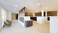 This generously sized single-floor space is designed as a modern communal facility, combining functional and social areas within a cohesive, contemporary aesthetic. The layout includes two hallways and a combined kitchen and living room area, all unified by light-toned stone tile flooring, recessed lighting, and crisp white walls throughout.

The hallways are bright and welcoming, featuring floor-to-ceiling windows along one corridor that invite abundant natural light and provide views of the surrounding outdoor landscape. Mid-century modern accent chairs are positioned along the windowed wall, creating casual seating nodes. A dedicated mail center is integrated into the hallway area, with rows of matte black mailboxes lining the walls alongside striking large-format artwork and dark wood slatted accent panels that add warmth and texture.

The combined living and lounge area is expansive and multi-functional, accommodating a billiards table, comfortable seating arrangements, and a curated gallery wall displaying illuminated artwork on dark wood paneling. A more intimate lounge nook features rich jewel-toned furnishings, a sculptural wall installation, and dark horizontal wood cladding, creating a vibrant contrast to the otherwise neutral palette. Double glass doors connect the living area to the exterior, further enhancing the sense of openness.