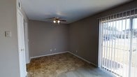 This single-floor space offers a straightforward and practical layout designed for everyday comfort. The main living area features earthy, stone-patterned tile flooring that transitions seamlessly to carpeted areas, providing both durability and warmth underfoot. The walls are finished in a neutral taupe tone, creating a calm and versatile backdrop that suits a variety of furnishing styles. A ceiling fan with an integrated light fixture is centrally mounted, offering both ambient lighting and year-round climate comfort. The textured popcorn ceiling adds a classic residential character to the space. A sliding glass door fitted with vertical blinds allows for natural light to filter into the room while providing direct access to the outdoors, offering views of the exterior surroundings. Electrical outlets are conveniently placed along the walls, and white baseboards provide a clean finishing detail throughout. The overall ambiance is modest and functional, with a neutral palette that makes the space easy to personalize. The single-floor layout ensures ease of navigation, making it a practical and accessible living environment.