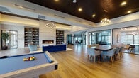 Located at 77 Gardnertown Road in Newburgh, NY, this single-story clubhouse is a thoughtfully designed amenity space that balances elegance with functionality. Entry is made through a dramatic arched glass vestibule, where soaring vaulted ceilings and large steel-framed glass doors flood the foyer with natural light, setting a refined tone throughout.

The expansive main lounge serves as the heart of the space, anchored by a floor-to-ceiling travertine-clad linear fireplace flanked by custom built-in shelving. Rich wood-paneled ceilings, warm hardwood floors, and sculptural globe chandeliers contribute to the sophisticated ambiance. Plush navy velvet seating and accent chairs create an inviting gathering area, while a billiards table offers a recreational focal point.

Adjacent to the lounge, a generous dining area accommodates group seating beneath stylish pendant lighting, with a bar-height counter providing additional seating options. The fully equipped kitchen features stainless steel appliances, a professional-grade range with a hood, mosaic tile backsplash, natural wood cabinetry, and a large center island with quartz countertops.

Large windows and glass doors throughout the space provide views and direct access to an outdoor pool area, enhancing connectivity to the exterior. Restroom facilities and a fitness center complete this well-appointed clubhouse.