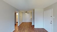 This single-floor residence offers a clean, functional layout designed for comfortable everyday living. The main living area is highlighted by warm-toned hardwood flooring that extends throughout, creating a cohesive and inviting atmosphere. Neutral gray-toned walls provide a versatile backdrop, complemented by white baseboards and trim that add a crisp, polished finish. A ceiling fan with a built-in light fixture is centrally mounted, offering both practical ventilation and ambient lighting for the space. The open layout allows for a natural flow between the living area and adjoining rooms. A doorway leads directly into a well-lit kitchen, which features white cabinetry and appears to benefit from ample natural light. Overhead HVAC vents are integrated into the ceiling, ensuring efficient climate control throughout. The space maintains a straightforward, move-in-ready character with minimal architectural ornamentation, placing emphasis on functionality and ease of use. The overall ambiance is bright and airy, making it a practical and comfortable living environment suitable for a variety of uses.