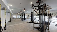 Fitness Center 2 is a thoughtfully designed, single-level facility that offers a comprehensive workout environment suited to a variety of fitness routines. The open floor plan is anchored by neutral-toned flooring with a subtle marbled texture, which lends the space a clean, contemporary aesthetic throughout.

The strength training area features a multi-function cable machine and a heavy punching bag, supported by floor-to-ceiling mirrors along one wall that visually expand the space and allow users to monitor their form. Ceiling fans are evenly distributed overhead, complementing the recessed lighting to maintain a comfortable atmosphere during workouts.

The cardio section is well-appointed with rowing machines and stationary cycling bikes positioned near expansive floor-to-ceiling windows. These large windows invite abundant natural light and provide open views of the surrounding exterior, creating an energizing environment for cardio training.

A dedicated functional fitness area includes a stability ball rack stocked with multiple sizes, a wall-mounted yoga mat, and additional accessories for stretching and core work. A flat-screen television is mounted on the wall, visible from multiple areas of the room.

Decorative circular wall accents add a modern design element, while structural columns help define distinct zones within the space. The overall ambiance is bright, airy, and professionally appointed, offering a well-rounded fitness experience.