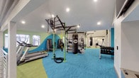 This fitness center is a thoughtfully designed, single-level space that seamlessly blends high-energy aesthetics with comprehensive workout functionality. The flooring makes an immediate visual impact, featuring a bold two-tone pattern of teal and lime green textured carpet that defines different workout zones throughout the space. Walls are accented with vibrant teal and green paint, large-scale athletic photography murals, and motivational imagery that reinforce an active atmosphere.

The functional training zone serves as the centerpiece, anchored by a green TKO power rig system complete with gymnastic rings, battle ropes, and a suspended heavy punching bag. Nearby, a wall-mounted dumbbell rack provides an organized free weights station. Cardio equipment, including treadmills and rowing machines, lines designated areas along the perimeter.

An adjacent zone features additional strength and cable machines, rounding out the equipment offerings. A built-in planter display with tile mosaic detailing and pendant lighting adds a design-forward touch near a wall-mounted flat-screen television. The space opens to a mezzanine-level overlook adorned with decorative globe pendant lights, offering views into the broader community area below. Recessed lighting throughout ensures the entire fitness center is well-illuminated.