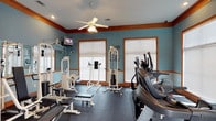 This fitness room offers a dedicated space for a comprehensive workout experience. The room is finished with teal-painted walls accented by warm wood crown molding and window trim, creating a visually appealing environment that balances energy with calm. A ceiling fan with integrated lighting is centrally mounted, ensuring air circulation and comfort during exercise sessions.

The space is outfitted with a variety of both cardio and strength training equipment. Cardio offerings include treadmills and elliptical machines positioned near the windows, allowing users to enjoy natural light while exercising. On the strength training side, the room features cable machines, weight benches, and additional resistance equipment, providing a well-rounded selection for full-body workouts.

The flooring consists of dark rubber matting, which provides cushioning, durability, and slip resistance—practical choices for a fitness environment. Multiple large windows fitted with white horizontal blinds allow ample natural light to fill the room while offering privacy control. A wall-mounted television adds an entertainment option during workouts. Overall, the space is thoughtfully designed to support a versatile and comfortable fitness routine.