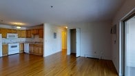 This single-floor unit offers a straightforward, functional layout centered around an open living area with warm hardwood flooring that extends throughout the space. The neutral white walls and standard-height ceilings create a clean, versatile backdrop suitable for a variety of furnishing styles.

The kitchen occupies one corner of the space and is separated from the main living area by a partial dividing wall. It features honey-toned wooden cabinetry providing ample storage, a double-basin sink positioned along the counter, and a compact, efficient footprint. An electrical panel is mounted on the adjoining wall.

Adjacent to the living area, a built-in closet with a bifold door provides convenient storage space, with a visible rod for hanging items. A doorway with a bright yellow-toned door leads to an additional area of the unit, suggesting a separate room or entryway beyond.

A baseboard heating unit runs along the far wall, indicating a reliable heating system suited to colder climates. The overall ambiance of the space is simple and practical, offering a blank canvas that can be easily personalized.