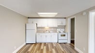 Located at 15025 Pine Meadows Dr in Fort Myers, Florida, this single-floor residence offers a clean, functional layout designed for comfortable everyday living. The open-concept living and kitchen area serves as the heart of the home, where warm-toned carpet in the living space transitions seamlessly to durable wood-look vinyl flooring in the kitchen. The kitchen is well-appointed with crisp white cabinetry offering ample storage, a single-basin sink, a white electric range, and a full-size refrigerator, all illuminated by a bright overhead skylight-style light fixture framed by a drop soffit ceiling detail. The living area is bathed in natural light from a front-facing window adjacent to the entry door, creating a welcoming atmosphere. The bedroom continues the warm carpet flooring and features a ceiling fan with light, a window with outdoor views, and a textured ceiling with crown molding details. The full bathroom is neatly finished with a white vanity and countertop, a gold-framed mirror, and a tub-shower combination. A separate utility room houses the water heater and electrical panel, adding practical functionality. Neutral wall tones and consistent white trim throughout give the space a cohesive, move-in-ready feel.