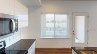 This single-floor space presents a clean, contemporary design highlighted by warm hardwood-style flooring that flows throughout the main living areas. The open-concept layout seamlessly connects the kitchen and living space, creating a sense of spaciousness and ease of movement. The kitchen is well-appointed with white cabinetry, dark granite countertops, stainless steel appliances including a range and microwave, and stylish pendant lighting that adds a modern touch overhead. A kitchen island with a stone-look surround provides additional prep and seating space.

The living area benefits from multiple large windows dressed with horizontal blinds, allowing generous natural light to fill the room while offering views of the surrounding neighborhood. A glass-paneled exterior door further enhances connectivity to the outdoors. The bedroom features plush carpeting and an impressive bank of floor-to-ceiling windows that frame expansive outdoor views, creating a bright and airy retreat. A hallway connects the main living spaces to additional rooms, including what appears to be a laundry area. Throughout, crisp white walls and clean architectural lines contribute to a fresh, move-in-ready ambiance.
