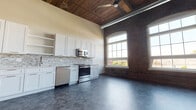 This single-floor loft-style unit masterfully combines historic industrial character with contemporary design. The open-plan main living and kitchen area immediately impresses with soaring wood-plank ceilings, exposed ductwork, and original brick walls punctuated by tall arched windows that flood the space with abundant natural light and offer outdoor views. Rustic wooden support columns add to the authentic character of the space.

The kitchen is well-appointed with crisp white shaker-style cabinetry, quartz countertops, a stone mosaic tile backsplash, and a full suite of stainless steel appliances including a range, microwave, and dishwasher. A ceiling fan overhead adds both function and style to the open living area.

The bedrooms are calm and neutral, featuring durable tile-style flooring, clean painted walls, and ample room for furnishings. A spacious walk-in closet with wire shelving systems provides generous storage.

The bathroom is elegantly finished with large-format tile flooring, a white vanity with ample cabinetry, and a fully tiled walk-in shower enclosed by a glass door. Throughout the unit, paneled interior doors with modern lever hardware and a consistent gray-toned tile flooring palette create a cohesive, polished aesthetic.