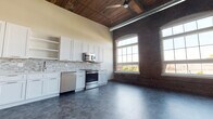 This single-floor loft-style unit masterfully combines historic industrial character with contemporary design. The open-plan main living and kitchen area immediately impresses with soaring wood-plank ceilings, exposed ductwork, and original brick walls punctuated by tall arched windows that flood the space with abundant natural light and offer outdoor views. Rustic wooden support columns add to the authentic character of the space.

The kitchen is well-appointed with crisp white shaker-style cabinetry, quartz countertops, a stone mosaic tile backsplash, and a full suite of stainless steel appliances including a range, microwave, and dishwasher. A ceiling fan overhead adds both function and style to the open living area.

The bedrooms are calm and neutral, featuring durable tile-style flooring, clean painted walls, and ample room for furnishings. A spacious walk-in closet with wire shelving systems provides generous storage.

The bathroom is elegantly finished with large-format tile flooring, a white vanity with ample cabinetry, and a fully tiled walk-in shower enclosed by a glass door. Throughout the unit, paneled interior doors with modern lever hardware and a consistent gray-toned tile flooring palette create a cohesive, polished aesthetic.