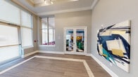 This single-floor amenity space is thoughtfully designed to balance energetic social areas with functional wellness facilities. The entrance foyer features wood-look tile flooring with a contrasting inlay border, crown molding, tall windows, and a set of glass French doors that offer a preview of the lively interior beyond. Bold abstract artwork accents the walls, establishing the space's vibrant aesthetic.

The main lounge area is expansive and light-filled, with large windows dressed in roller shades and patterned curtains. Striking large-format hexagonal tiles in a multi-tone palette span the floor throughout this zone. Seating arrangements include a vivid magenta sectional sofa paired with colorful abstract art on soaring walls, emerald green lounge chairs, and a dedicated conversation nook with teal barrel chairs surrounding a round coffee table.

The game and entertainment zone features a bold teal accent ceiling, a pool table, bar-height seating along a yellow-tiled wall, mounted televisions, and pendant lighting, creating a lively social hub. Colorful artwork continues throughout this area.

The fitness center is well-equipped with strength machines, free weights, battle ropes, and functional training equipment. A checkered carpet tile floor, large mirrors, recessed lighting, ceiling fans, and ample natural light from multiple windows create an energizing workout environment. A hydration station with a dual water fountain is conveniently accessible nearby.