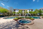 Located at 6929 Commerce Circle in Baton Rouge, Louisiana, this outdoor amenity space offers a resort-inspired retreat designed for relaxation and leisure. At the heart of the space is a generously sized freeform swimming pool with stone-edged steps leading into the water, complemented by an active water fountain feature that adds a tranquil ambiance to the surroundings. Adjacent to the pool sits a circular spa, finished with natural stone detailing that ties seamlessly into the overall aesthetic.

The pool deck is thoughtfully appointed with an array of modern lounge chairs and shade umbrellas, providing ample space for sunbathing and unwinding. A wooden pergola fitted with string lights anchors one side of the courtyard and houses an outdoor grilling station, making it ideal for gatherings. Additional seating areas with patio furniture and large umbrellas are scattered throughout, offering shaded retreats beneath a canopy of mature trees.

The entire area is enclosed by wrought iron fencing with gated access, ensuring privacy and security. Lush, manicured landscaping—including flowering shrubs, ornamental grasses, and established trees—frames the space beautifully against the backdrop of the multi-story stucco building. Under clear blue Louisiana skies, this outdoor space delivers a polished and welcoming environment.