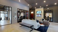 This single-floor space showcases a sleek, contemporary design that exudes a polished and welcoming ambiance. The lounge area is anchored by a striking natural stone accent wall, which serves as a focal point and frames a pair of mounted flat-screen displays — ideal for entertainment or informational use. Recessed lighting is thoughtfully distributed throughout the curved ceiling, casting a warm and even glow across the room. The furnishings are modern and sophisticated, featuring a clean-lined white sofa, a patterned upholstered bench with chrome legs, and coordinating lounge chairs arranged to encourage conversation and comfort. A navy area rug defines the seating zone and adds depth to the light hardwood flooring beneath. Adjacent to the main lounge, a glass-enclosed room with dark metal framing offers a distinct, semi-private area, adding visual interest and functional versatility to the layout. Decorative touches such as a tall arc floor lamp, colorful accent pieces, and wall art contribute to the refined yet approachable atmosphere. The overall design balances style and functionality, making it a well-appointed space suited for relaxation or social gatherings.