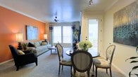 This single-floor residence offers an open-concept layout that integrates the dining and living areas into one cohesive, functional space. The living room is anchored by a bold orange accent wall, adding warmth and personality to the room, while a ceiling fan overhead ensures year-round comfort. Large windows dressed with patterned curtains flood the space with abundant natural light, creating a bright and welcoming atmosphere. The living area provides ample room for a full sofa arrangement, complemented by a coffee table and soft carpeted flooring that extends throughout.

Adjacent to the living room, the dining area accommodates a round table with elegantly styled upholstered chairs, making it well-suited for both casual meals and entertaining. Crown molding adds a refined architectural detail to the space. A glass-paneled door leads to an outdoor area, further enhancing the sense of openness and allowing natural light to pour in from multiple directions. Track lighting near the entry provides focused illumination, while fresh floral arrangements add a lively, curated touch to the overall ambiance of the space.