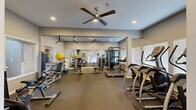 Located in Lynnwood, Washington, this well-appointed fitness and wellness facility spans a single floor and is divided into three distinct areas designed to support a comprehensive approach to health and relaxation. The main gym area is generously sized, featuring durable rubber flooring, recessed lighting, and dual ceiling fans that ensure a comfortable workout environment. Large windows fitted with blinds allow ample natural light to fill the space. The gym is fully equipped with a wide variety of strength and cardio equipment, including treadmills, elliptical machines, a cable machine, free weights, medicine balls, stability balls, weight benches, and a stationary bike. Full-length mirrors line one wall, enhancing the sense of space and allowing users to monitor their form. A wall-mounted television and a pull-up bar add to the functionality of the area. An adjoining cardio-focused zone offers additional treadmills and elliptical trainers positioned alongside a bank of windows. The third area is a warm, intimate indoor hot tub room finished with rich wood-paneled walls and ceiling, creating a cozy, spa-like atmosphere. The hot tub is tiled in blue and framed by brick surround, with seating nearby for relaxation. Together, these spaces provide a complete fitness and recovery amenity.