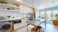 This single-floor unit presents a cohesive modern design characterized by light wood flooring, neutral tones, and clean lines throughout. The kitchen serves as a functional centerpiece, outfitted with flat-front cabinetry in white and warm wood tones, a subway tile backsplash, stainless steel appliances including a gas range and full-size refrigerator, and an under-counter microwave. Open shelving adds display space, while a freestanding stainless steel island with woven basket storage below provides additional prep space and casual seating. Natural light enters through a window overlooking the surrounding neighborhood. The bedroom is compact and efficiently arranged, featuring a window framing an urban cityscape that includes a notable domed building on the horizon, contributing a distinctive character to the room. The bathroom is finished in a clean, minimalist style with light wood flooring, a white vanity with an undermount sink and large mirror, and a walk-in shower enclosed by a fabric curtain. The overall layout flows logically between spaces, and the consistent use of light materials and neutral finishes gives the unit a bright, airy ambiance throughout.