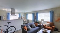 This single-floor space showcases an open-concept layout that seamlessly connects the living, dining, and kitchen areas into one cohesive environment. The living area is anchored by a charcoal gray sofa accented with burnt orange and patterned throw pillows, complemented by a matching orange armchair that adds a warm pop of color. Hardwood-style flooring runs throughout, providing a clean, modern foundation.

Multiple windows fitted with white horizontal blinds and bright blue curtains line the walls, allowing generous amounts of natural light to fill the space while adding a lively visual contrast to the neutral gray walls. A track light fixture overhead ensures consistent illumination throughout the open area.

The dining area, visible beyond the living space, features a modest dining table with seating, positioned near a pass-through opening that connects to the kitchen. The kitchen is partially visible and includes overhead cabinetry and countertop appliances. The overall ambiance is casual yet well-styled, with a balanced mix of cool and warm tones creating an inviting, comfortable atmosphere throughout.