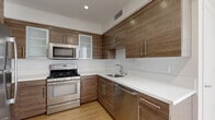 This single-floor unit offers five bedrooms, one bathroom, a kitchen, and a closet, all thoughtfully arranged within a compact and efficiently designed floor plan. Light wood flooring runs continuously throughout, creating a warm, cohesive aesthetic that unifies every room.

The kitchen stands out as a highlight of the space, showcasing rich wood-grain cabinetry with sleek bar-pull hardware, white quartz countertops, and a full suite of stainless steel appliances including a gas range, built-in microwave, and dishwasher. Frosted glass cabinet inserts add a refined touch, while recessed lighting and clean white walls keep the space bright and functional.

The bedrooms are bright and minimalist, each featuring white walls, light wood flooring, and natural light from large windows that offer views of surrounding greenery and neighboring architecture. The generously sized primary bedroom includes an open closet with built-in shelving and direct sightlines to the kitchen. Additional bedrooms are well-proportioned and flooded with natural light.

A clean hallway with crisp white walls and warm flooring connects the rooms, maintaining an open, airy feel throughout. The bathroom is neatly finished with white subway tile, a glass-enclosed shower-tub combination, chrome fixtures, and tile flooring, offering a clean and practical design. Overall, the space presents a modern, light-filled environment with consistent finishes throughout.