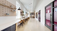 Located on East Colonial Drive in Orlando, Florida, this single-floor creative studio offers a generously sized, thoughtfully designed workspace tailored for artistic endeavors. The open layout is anchored by polished concrete floors throughout the main corridors and gathering areas, transitioning to paint-splattered concrete in the dedicated art-making zones — a testament to the space's vibrant creative activity.

The main corridor runs the length of the studio, lined with continuous counter-height worktables, adjustable drafting stools, and pegboard wall systems mounted beneath a row of large windows that flood the space with natural light. Industrial-style pendant lights hang overhead, complementing the exposed ductwork and high ceilings that lend the space an airy, warehouse-inspired character.

Separate rooms branch off the main corridor, enclosed by sleek black-framed glass partition walls and doors, allowing visual connectivity while defining distinct functional zones. One room is outfitted with floor-to-ceiling built-in shelving in light wood, stocked with art supplies, paper rolls, and storage drawers — ideal for organized, multi-medium creation. A utility sink station provides practical support for hands-on work.

The entry area features a striking large-scale mixed-media artwork displayed on a white accent wall, setting a gallery-like tone. A vibrant pink-accented room adds a bold pop of color, visible through the glass partitions. The overall ambiance is clean, modern, and purposefully designed to inspire creativity at every turn.